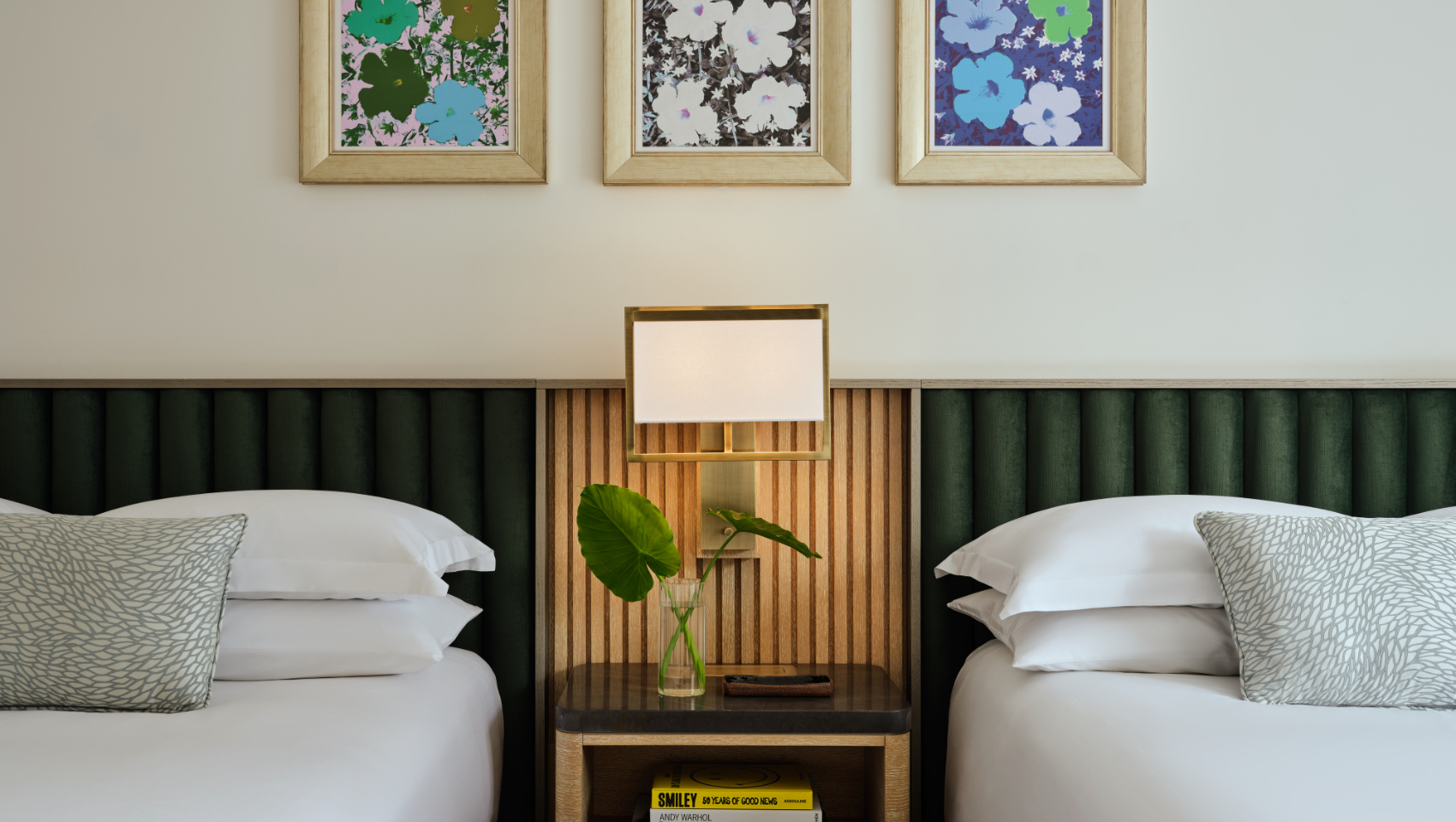 A hotel room with two beds, green headboards, a bedside table with a lamp, and three framed pop art flower prints